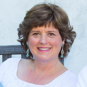 May include: A woman with brown hair and brown eyes smiles at the camera. She wears turquoise and silver earrings and a white top. The background is a light-colored wall.