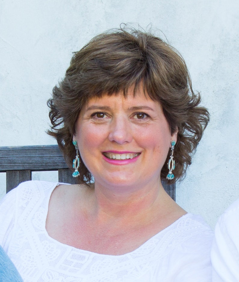 May include: A woman with brown hair and brown eyes smiles at the camera. She wears turquoise and silver earrings and a white top. The background is a light-colored wall.