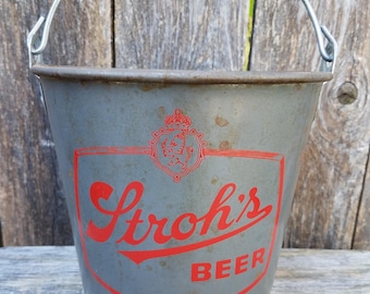Könnte beinhalten: Ein grauer Metalleimer mit einem silbernen Drahtgriff. Die Vorderseite zeigt ein rotes grafisches Logo mit einer Krone, einem Löwenemblem und dem Text Stroh's BEER in einer fetten, stilisierten Schrift. Die Metalloberfläche weist leichte Oxidationsspuren auf.