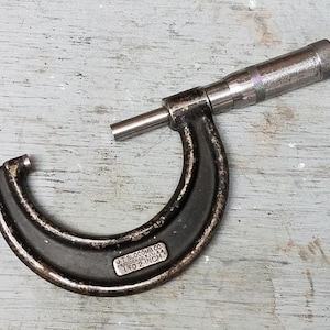 May include: A vintage black and silver micrometer measuring tool. The tool is marked "J.T. Slocomb Co. Providence R.I. U.S.A. 1 to 2 inch". The micrometer is a U-shaped tool with a rotating barrel and a measuring rod.