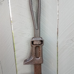 May include: A heavy-duty, dark brown cast iron pipe wrench featuring a long, slender handle with an integrated oval hanging loop at the top, a central adjustable sliding jaw mechanism, and a fixed bottom jaw with a textured, rugged surface.