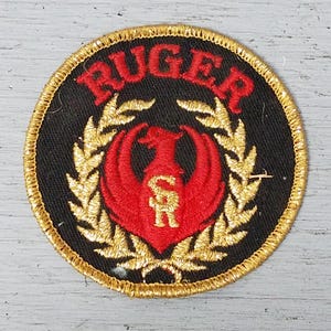May include: A round, black patch with the word "RUGER" in red arched across the top. A red phoenix-like bird is centered, with gold laurel branches and a gold border.