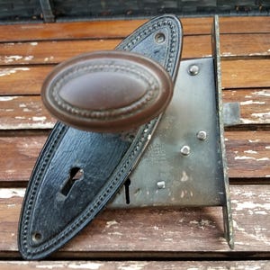 May include: A vintage mortise door lock set featuring a dark brown oval knob attached to an elongated black metal backplate with a beaded edge and a keyhole. The internal mechanism is a rectangular metal box with visible rivets and a latch.