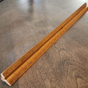 Puede incluir: Una regla de arquitecto vintage de madera, con marcas en pulgadas y fracciones. La regla es de color marrón claro con marcas negras y la palabra "POST" impresa. La escala mide aproximadamente 30 cm de largo.