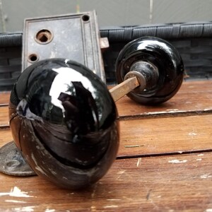 May include: A set of two round, glossy black door knobs with a metallic spindle connecting them, positioned in front of a rectangular, dark-colored metal door latch mechanism with visible screw holes and a central square opening for the spindle.