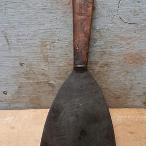 May include: A metal putty knife with a wide, triangular, dark grey blade showing signs of wear and surface oxidation. The tool features a long, rectangular wooden handle secured with two visible metal rivets near the top and bottom.