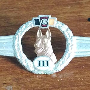 May include: A silver-coloured metal badge featuring a German Shepherd dog head within a wreath. The badge has a stylized wing design on either side and a small rectangular emblem at the top with coloured details. The bottom has the Roman numeral III.