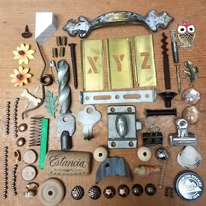 May include: A collection of vintage hardware and decorative items, including a metal handle, brass stencils with "XYZ" cutouts, keys, a cork, and a bottle cap. Various small metal and wooden pieces are also present, arranged on a wooden surface.