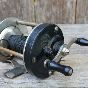 Può includere: Un mulinello da pesca vintage caratterizzato da una piastra laterale circolare nera con il testo in rilievo Acme, un telaio in metallo argentato e una bobina avvolta con lenza nera. Il mulinello include una manovella doppia con pomelli neri.