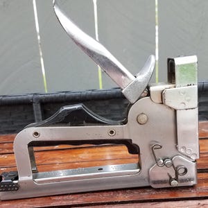 May include: A vintage silver and black staple gun. The tool has a curved handle, a lever, and a staple chamber. It is designed for heavy-duty use and is likely used for upholstery or construction projects.