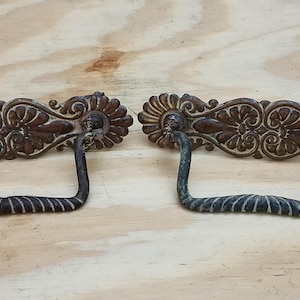 May include: Two antique bronze drawer pulls with ornate floral and leaf designs. The pulls have a dark brown finish with a twisted handle. The hardware is set against a light wood surface.