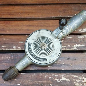 Może przedstawiać: Wiertarka ręczna vintage marki Craftsman z korpusem z metalu w kolorze srebrnym z widocznymi przetarciami farby, okrągłą obudową przekładni z koncentrycznymi pierścieniami, czarną korbą i ciemnobrązowym uchwytem wiertarskim.