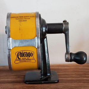 Puede incluir: Un sacapuntas manual vintage con un cuerpo cilíndrico de color amarillo brillante y una base y manivela de hierro fundido negro. La carcasa amarilla presenta texto negro que dice Carson Pirie Scott & Co. Stationery Section Chicago y Chicago Made By Automatic Pencil Sharpener Co. Chicago, U.S.A.