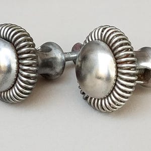 May include: Two silver-toned metal cabinet knobs featuring a smooth, rounded center dome surrounded by a textured, coiled wire border. Each knob includes a short, threaded metal post and a flat circular base for mounting to a drawer or door surface.