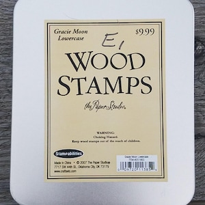 Pode incluir: Uma lata retangular branca contendo carimbos de madeira. O rótulo diz "Gracie Moon Lowercase WOOD STAMPS" com o logotipo Paper Studio. O preço é de $9,99. Um aviso sobre perigo de asfixia também é impresso no rótulo.