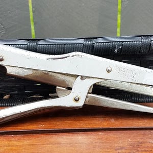 May include: A vintage Bostitch Model P3 stapler, made of silver-colored metal. The stapler is resting on a wooden surface, with a black woven background. The text "BOSTITCH MODEL P3" is visible on the side of the stapler.