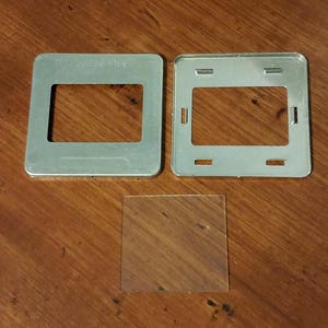 May include: Two silver metal frames with a clear glass insert. The frames have a rectangular shape with a cutout in the center. The frames are on a wooden surface.