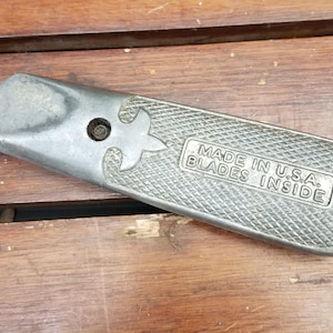 May include: A silver-toned metal utility knife with a textured diamond-patterned grip. The handle features a fleur-de-lis emblem and a rectangular plate embossed with the text MADE IN U.S.A. BLADES INSIDE. The tool has a tapered end.
