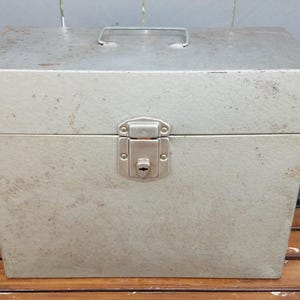 May include: A vintage metal storage box in a light gray color. The box has a rectangular shape with a hinged lid and a metal handle. A metal latch secures the lid. The box appears to be aged with some wear and tear.