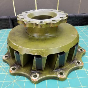 以下が含まれることがあります： オリーブグリーンの金属製機械ハブ部品。12個のボルト穴がある円形のトップフランジ、表面にひび割れが見られる中央の円筒形ボディ、8個の取り付けポイントと黒いゴム製インサートを備えた下部ベースで構成されています。