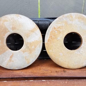 May include: Two circular stone discs with a central hole, featuring a mottled surface with shades of beige, cream, and light brown. The discs have a smooth, flat appearance and are positioned side by side, displaying natural colour variations.