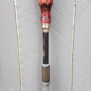 May include: A vintage push drill featuring a fluted, dark red wooden handle, a smooth black metal shaft, and a textured silver-colored metal collar with a flat-head bit at the bottom. The tool exhibits signs of wear and age on the handle and shaft.