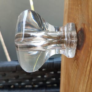 May include: A clear, faceted glass cabinet knob with a rounded, bulbous head and a square base, attached to a light brown wooden surface. The interior of the knob shows a metallic screw mechanism visible through the transparent glass material.