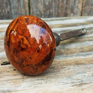 May include: A round door knob with a glossy, marbled finish in shades of deep amber, brown, and tan. The knob features a smooth, reflective surface and is attached to a dark, metallic square-shaped spindle extending from the back.