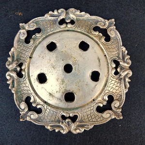 May include: A circular silver-toned metal plate featuring an ornate, scalloped border with embossed scale patterns and scrollwork. The center disc contains five evenly spaced circular cutouts surrounding a single, smaller central hole.