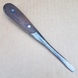 Könnte beinhalten: Ein Schlitzschraubendreher mit einem dunkelbraunen Holzgriff, der mit zwei silberfarbenen Metallnieten befestigt ist. Das Werkzeug hat einen langen, konischen Stahlschaft, der in einer flachen, rechteckigen Spitze für Schlitzschrauben endet.