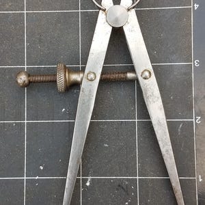 Puede incluir: Calibrador de metal vintage con un asa circular y puntas afiladas. La herramienta tiene un mecanismo de ajuste de tornillo y está hecha de metal de color plateado. El calibrador está colocado sobre una superficie de cuadrícula negra.