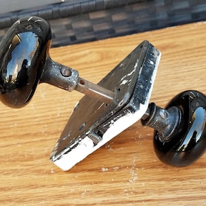 Peut inclure: Deux poignées de porte noires antiques avec une plaque métallique. Les boutons sont ovales et brillants, reflétant l'environnement. La plaque métallique est rectangulaire avec de la peinture blanche sur les bords. La quincaillerie est posée sur une surface en bois.