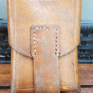 May include: A rectangular tan leather pouch featuring a vertical strap closure with visible light-coloured stitching. The leather has a weathered, textured surface with natural variations in colour and tone, showing a structured, boxy design.