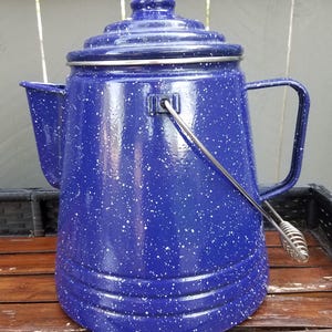 May include: A blue speckled enamelware coffee pot with a lid, handle, and spout. The pot has a silver handle and a classic design, perfect for camping or outdoor use. The pot is approximately 10 inches tall.