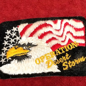 Pode incluir: Uma placa de madeira artesanal com o texto "Bem-vindo à nossa casa" em estilo manuscrito. A placa é pintada de branco com um acabamento desgastado e uma borda preta. Um pequeno coração decorativo está preso na parte inferior.