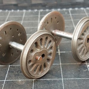 Puede incluir: Primer plano de dos juegos de ruedas de tren modelo antiguas. Las ruedas son de metal con un aspecto desgastado y envejecido. Cada juego incluye dos ruedas conectadas por un eje metálico. Las ruedas tienen patrones detallados de radios y remaches.