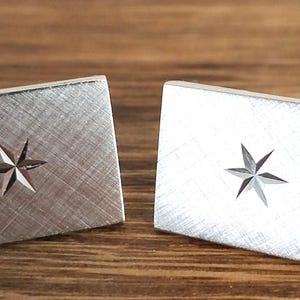 May include: A pair of rectangular silver cufflinks with a brushed metal finish. Each cufflink features a six-pointed star design in the center. The cufflinks are displayed on a wooden surface.