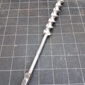 May include: A vintage metal auger drill bit with a spiral fluted design. The drill bit is silver-colored and has a pointed tip. The tool is designed for boring holes in wood. The background is a dark surface with a grid pattern.
