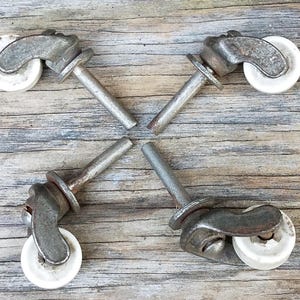 May include: Four vintage furniture casters featuring weathered gray metal brackets and white plastic wheels. Each caster includes a straight metal stem for mounting, showing signs of age and oxidation on the metal surfaces and hardware components.