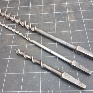 May include: Three vintage auger drill bits of varying sizes, arranged diagonally. The bits are made of metal, with a spiral design for wood boring. The drill bits have a dark, aged appearance, suggesting they are antique tools.