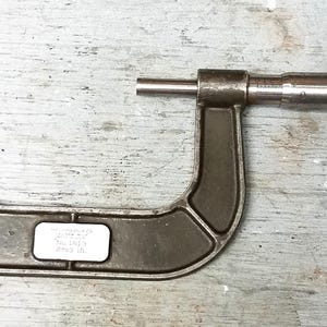 May include: A vintage, dark gray micrometer with a textured handle and a U-shaped frame. The tool, marked "2 in," is designed for precise measurements. It rests on a weathered, light gray wooden surface, showcasing its industrial design.