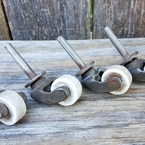 May include: A set of four vintage furniture casters, each featuring a dark, weathered metal mounting stem and bracket paired with a smooth, off-white ceramic wheel. The hardware displays signs of age and oxidation on the metal components.