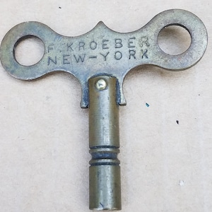 May include: A brass clock winding key featuring a T-shaped handle with two circular finger holes. The text F. KROEBER NEW-YORK is embossed on the front of the handle. The cylindrical shaft has two horizontal grooves and a small metal rivet.