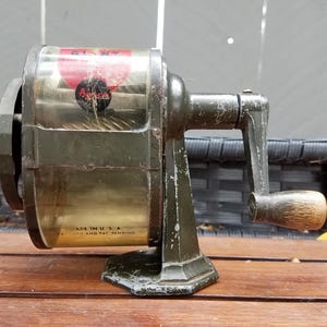 Puede incluir: Un sacapuntas manual vintage de color verde oliva con un depósito de virutas de plástico transparente. El dispositivo cuenta con una manivela con mango de madera, una etiqueta roja y negra que dice GIANT y el texto MADE IN U.S.A. en la base.