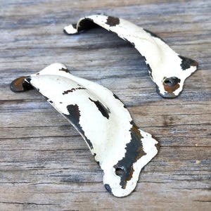 May include: Two curved metal drawer pulls featuring a distressed white enamel finish with visible dark brown rust patches. Each handle has a semi-circular grip and flat ends with pre-drilled screw holes for mounting to furniture or cabinets.