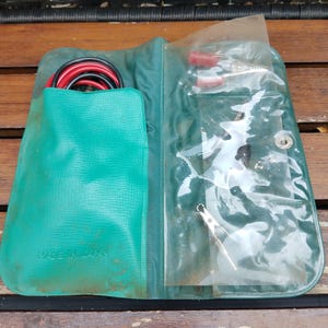May include: A green vinyl folding case containing automotive jumper cables with red and black insulated handles. The left side holds the coiled cables, while the right side features a clear plastic pocket holding metal clamps. Made in Japan.