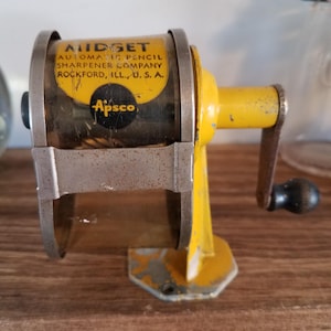 Pode incluir: Um apontador de lápis vintage amarelo e prateado com o texto "Budget Automatic Pencil Sharpener Company Rockford, ILL., U.S.A." e o logótipo "Apsco". O apontador tem uma manivela e um recipiente transparente.