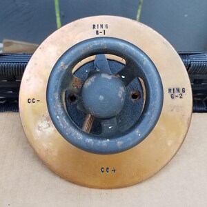 Könnte beinhalten: Eine kreisförmige, messingfarbene Metallplatte mit einem zentralen schwarzen Metallhandrad mit vier Speichen. Die Platte ist mit den Texten RING G-1, RING G-2, CC- und CC+ beschriftet. Das mittlere Rad weist Oxidationsspuren auf.