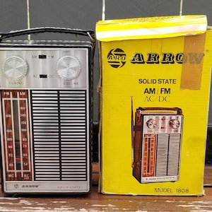 May include: A vintage Arrow solid state AM/FM radio with a silver face, tuning and volume knobs, and a black carrying handle, positioned next to its original yellow cardboard box featuring the Arrow logo and model 1808 text.