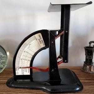 May include: A vintage black postal scale with a curved dial and a small platform. The dial reads "Ideal Postal Scale" and has weight measurements. A red lever is visible. The scale is on a wooden surface.